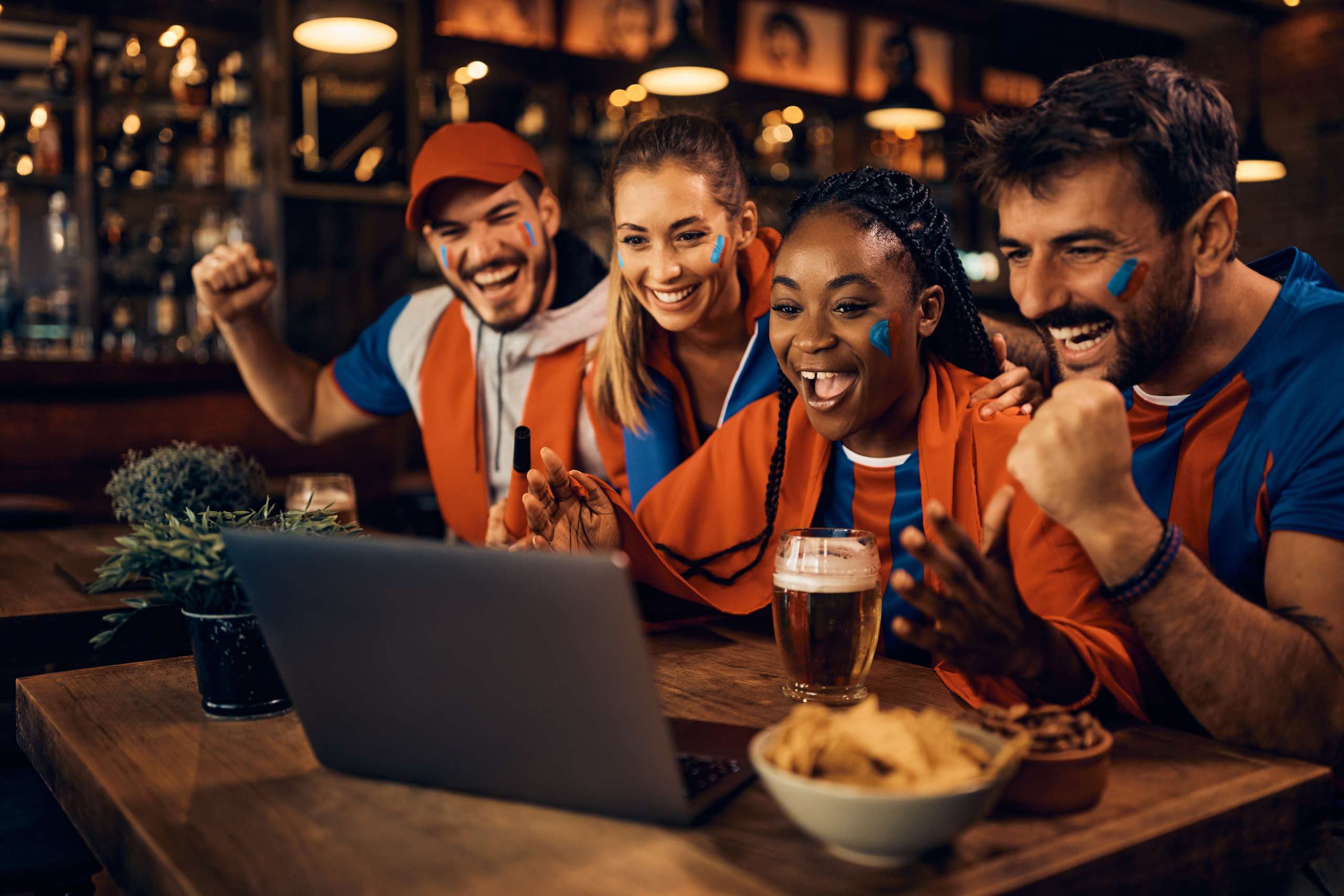 Group of cheerful soccer fans celebrating favorite team's victory while watching game on laptop in pub.